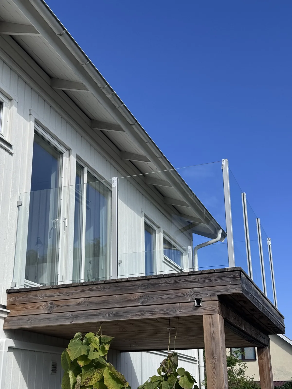 Glasräcke med metallstolpar längs en träbalkong vid ett vitt hus